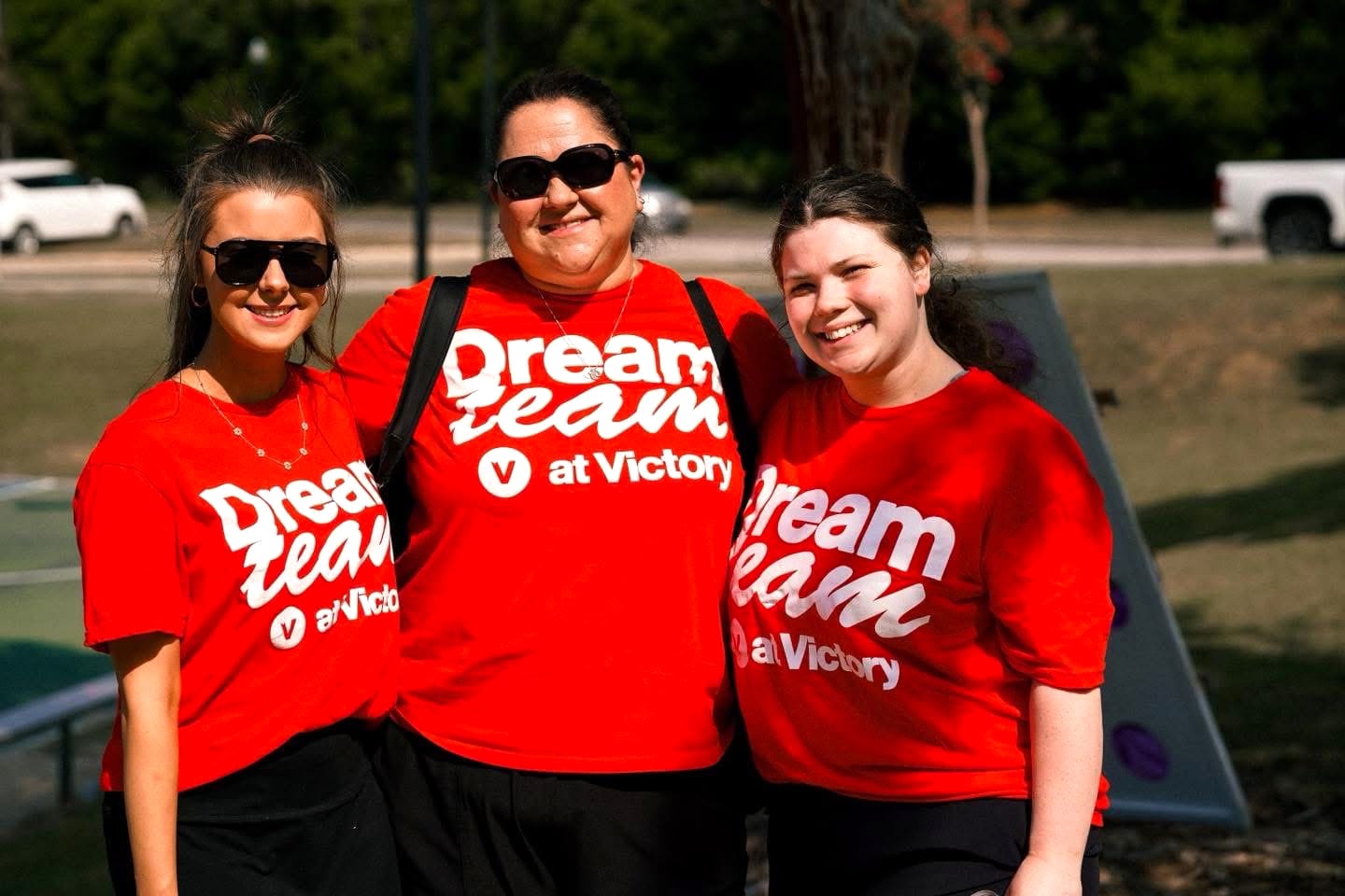 Three Dream Team members in red Dream Team shirts smiling together in the sunshine at an outdoor serve day